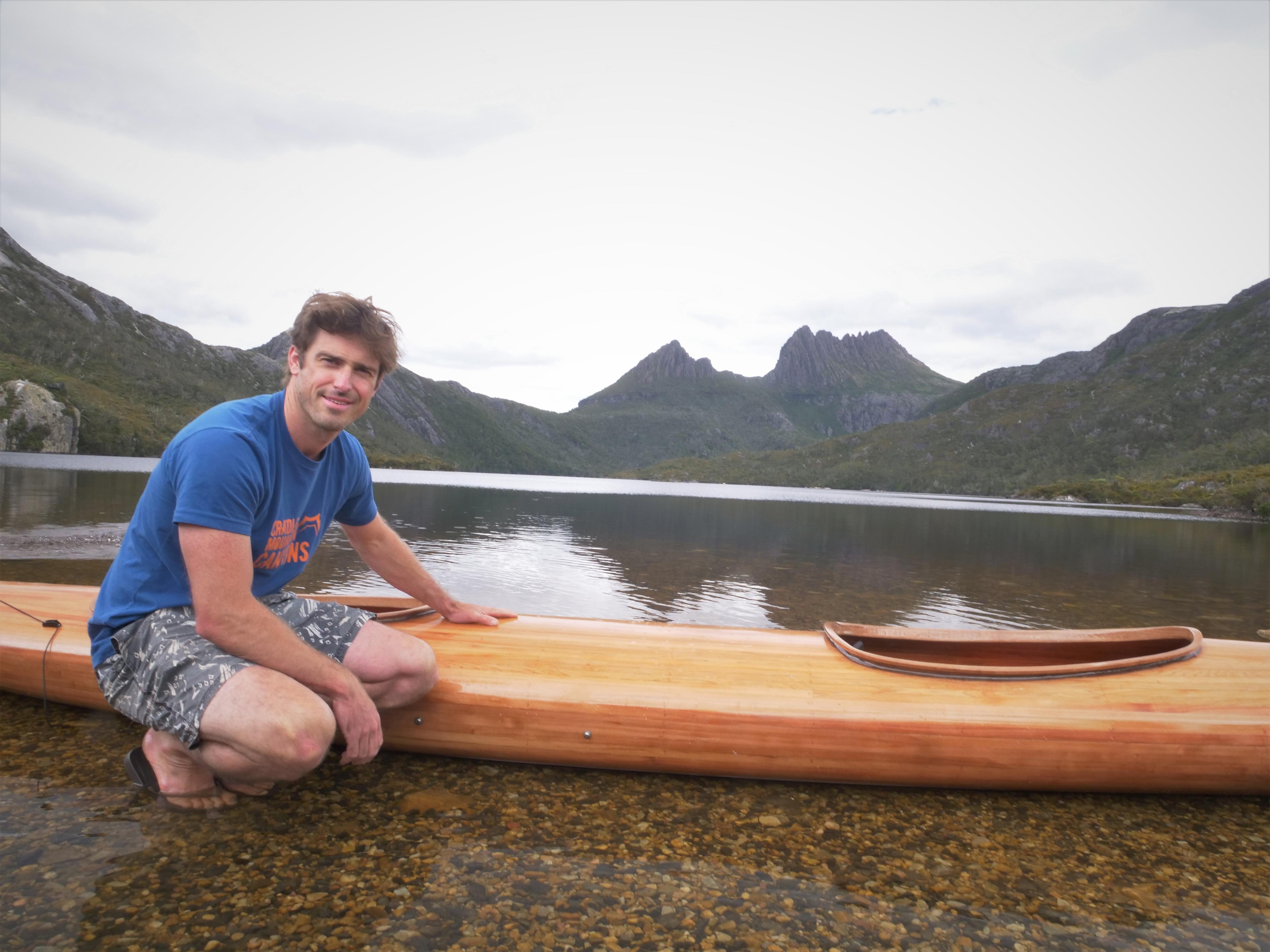 A man in shorts and tee shirt crouched by the edge of a lake, with his hand on a timber kayak and mountains behind.