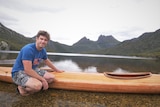 A man in shorts and tee shirt crouched by the edge of a lake, with his hand on a timber kayak and mountains behind.