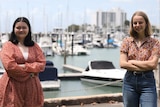 Two young woman stand at a marina with their arms crossed on a bright day.
