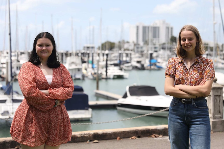 Two young woman stand at a marina with their arms crossed on a bright day.