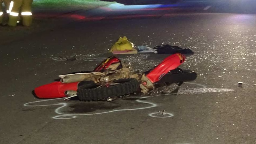 The shattered remains of a dirt bike lie on the road, as emergency services officers stand to one side.