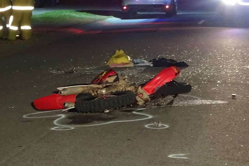 The shattered remains of a dirt bike lie on the road, as emergency services officers stand to one side.