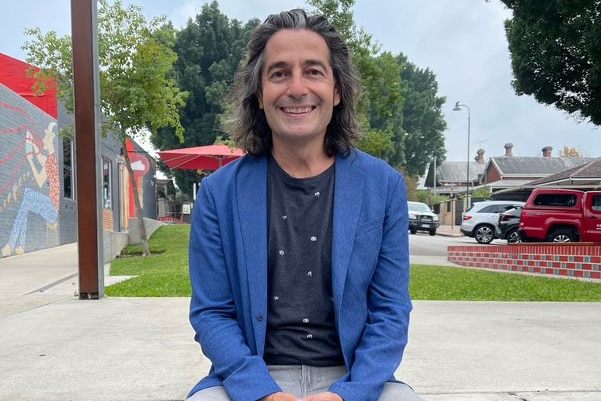 A man sits outdoors wearing a blue jacket. He has shoulder length dark hair.