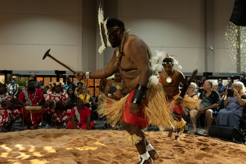 Two men in traditional Torres Strait Islander costume dance inside an art gallery, before a crowd.