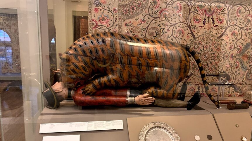 An automaton shaped like a tiger mauling a man sits in a glass cabinet alongside some guns and an embroidered hanging.