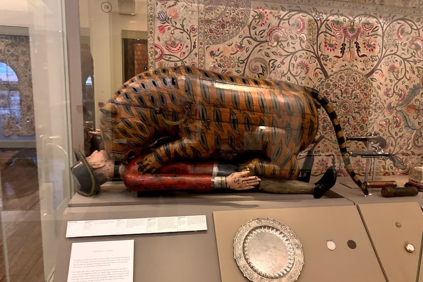 An automaton shaped like a tiger mauling a man sits in a glass cabinet alongside some guns and an embroidered hanging.