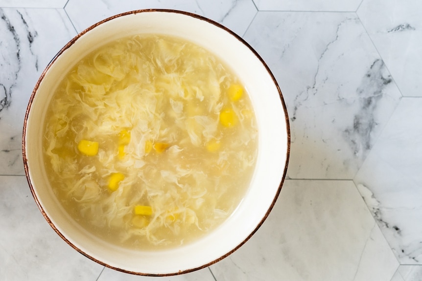 An aerial shot of a white bowl filled with clear broth, with corn kernels and chicken floating in it
