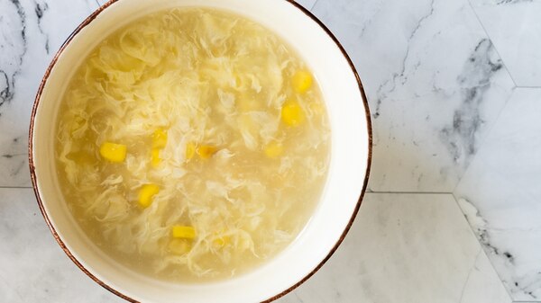 An aerial shot of a white bowl filled with clear broth, with corn kernels and chicken floating in it
