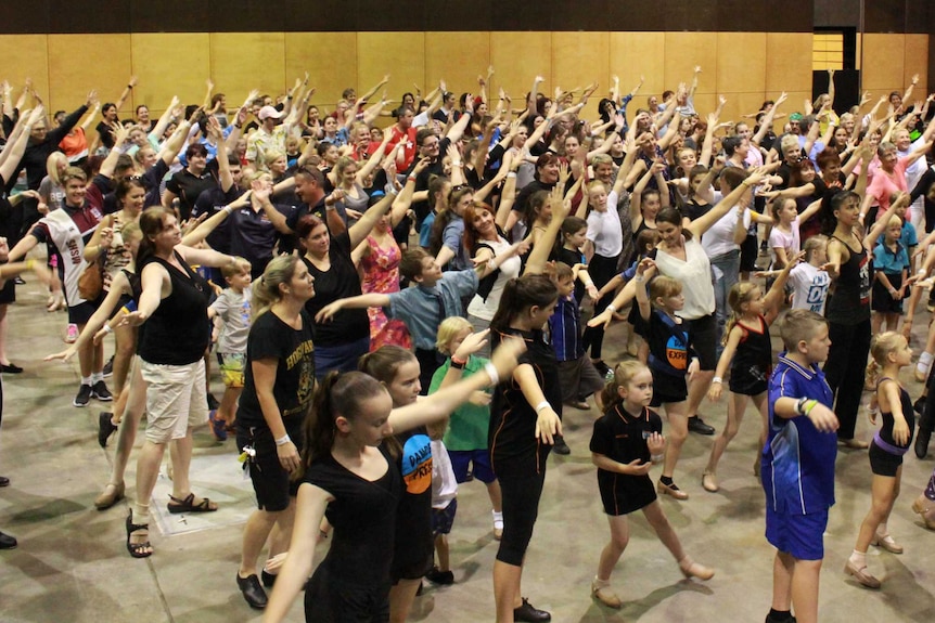 A large group of people raise their left hands in the air as part of a tap dancing move in a big tap dance class.