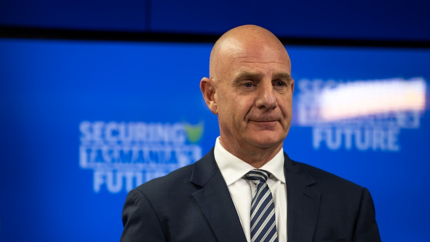 A bald man in a suit smiles in front of a blue screen 