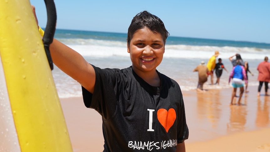 An Indigenous boy stands at the beach smiling, with the ocean behind him.