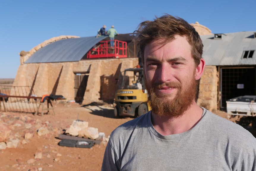 A man with short brown hair wearing a grey shirt stands in front of a woolshed while two men stand on the roof.