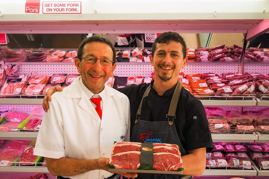 Two butchers stand in front of of a shelve full of meat while holding a rump stake