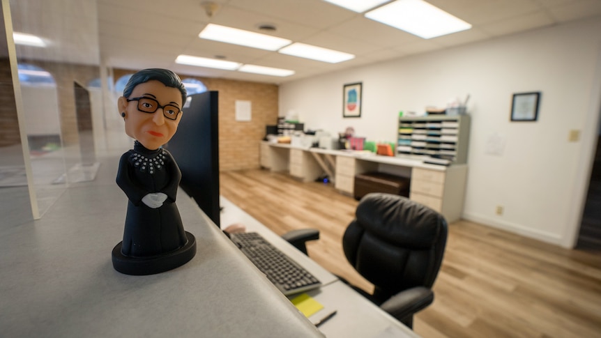 A Ruth Bader Ginsburg bobblehead sits atop a reception desk, an empty chair tucked in