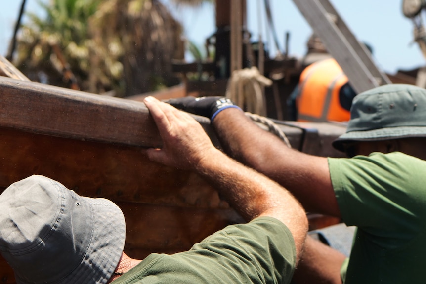 Two men in green t-shirts and bucket hats working on the side of a wooden boat.