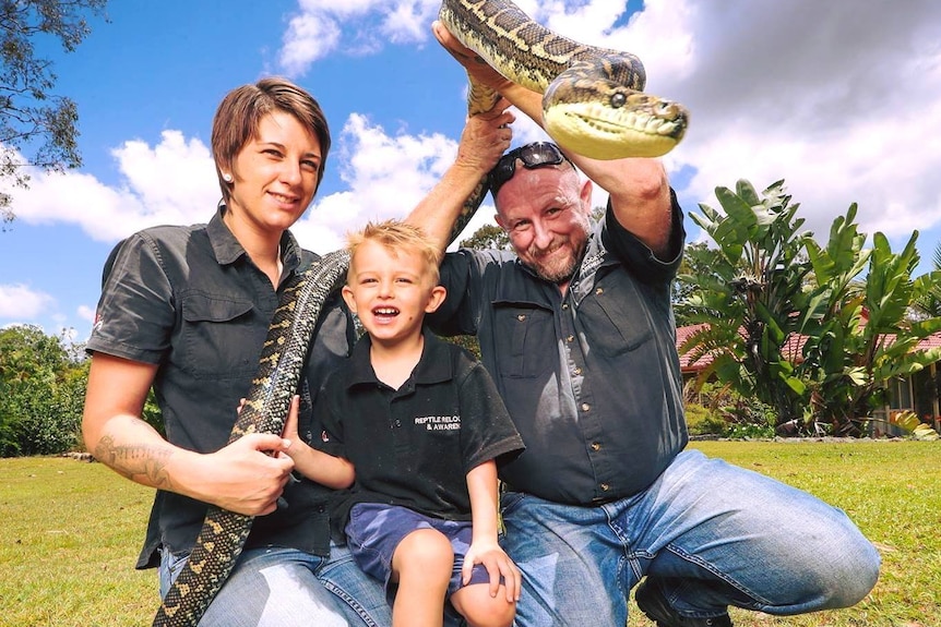 Man, woman and child kneeling on ground with man holding up a large python.