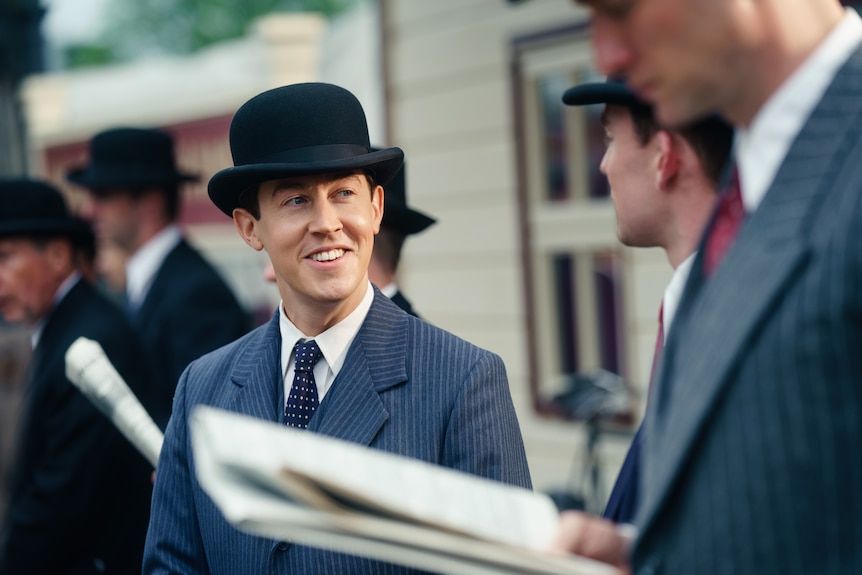 A 30-something man wearing a 50s-style suit and bowler hat smiles brightly, while standing at an old train station