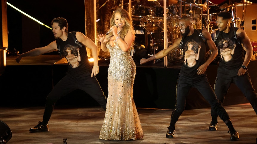 Mariah Carey sings on stage with three back up dancers, all wearing T-shirts with her face on them. 