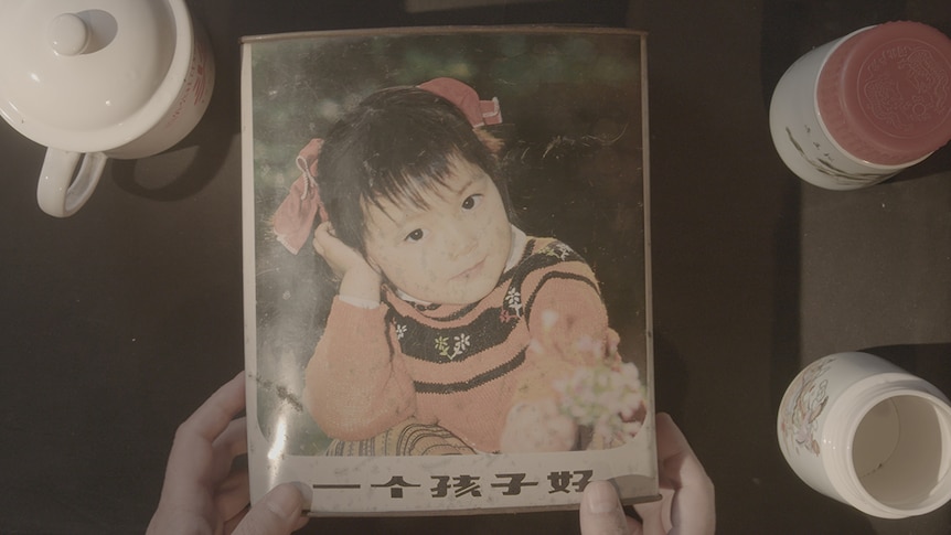 Colour still top down view of hands holding a picture of a child over table in 2019 documentary One Child Nation.