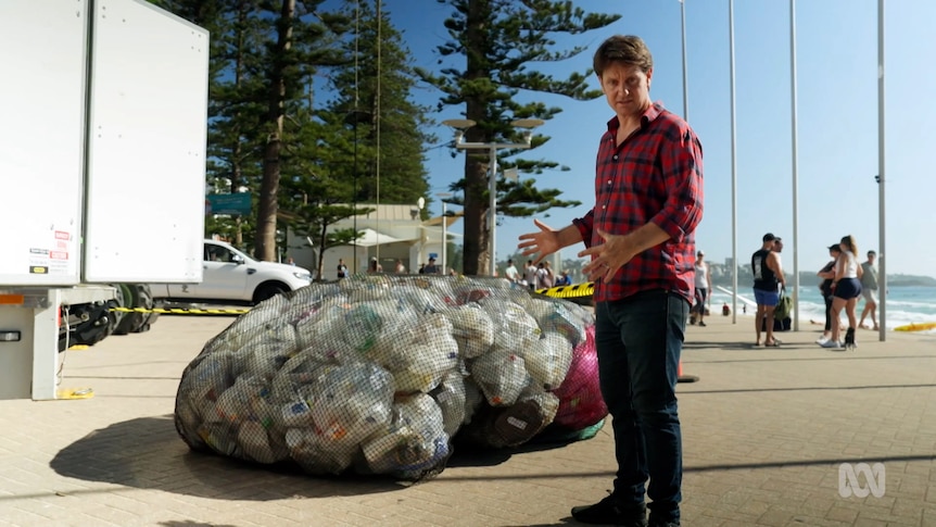 Craig Reucassel stands in front of a big fishing net full of waste plastic