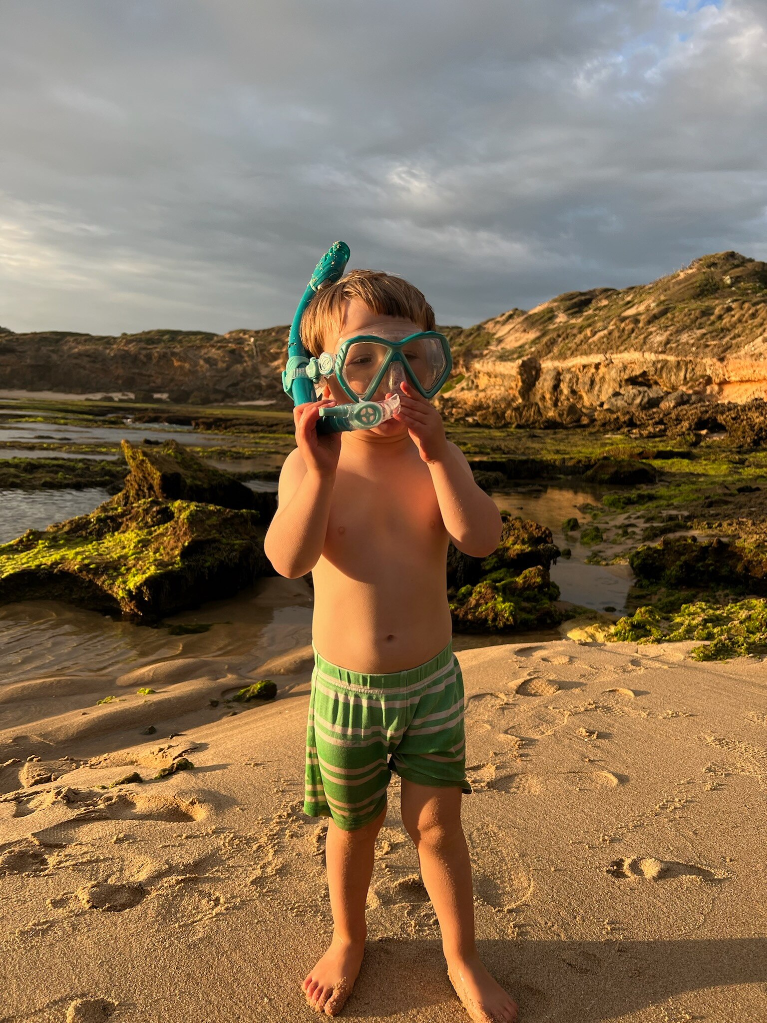 A small boy in bathers and a blue snorkel in front of some rocks.