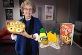 A middle-aged woman with blonde curly hair holds a round dish with a pineapple slice in it.