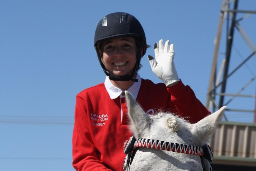 A young woman sitting on a horse in riding gear, waving and smiling.