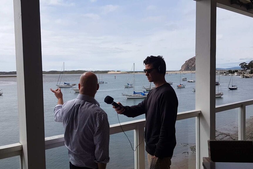 A man in a black sweater interviewing another man by the seaside.