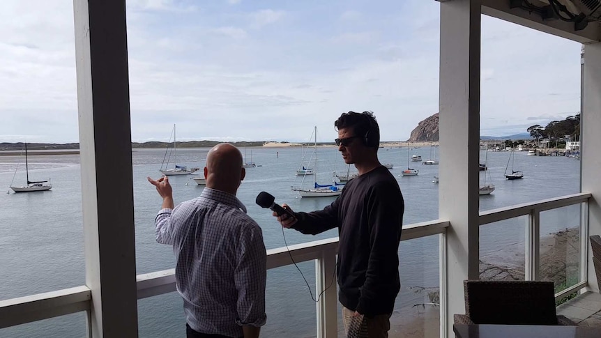 A man in a black sweater interviewing another man by the seaside.