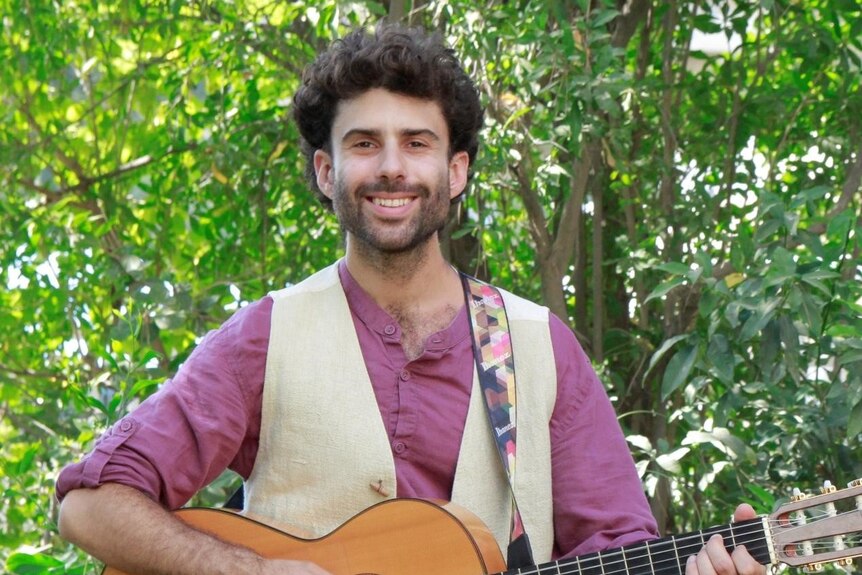 Man holding a guitar outdoors with trees in the background, smiling