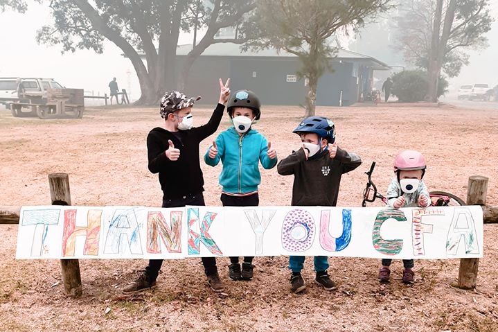 Three young children wearing air filter masks and bike helmets pose in front of a hand drawn thank you sign.