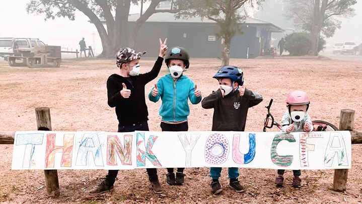 Three young children wearing air filter masks and bike helmets pose in front of a hand drawn thank you sign.