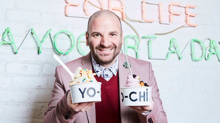 George Calombaris smiles at the camera holding two punnets of frozen yoghurt infront of a white wall with writing in lights.