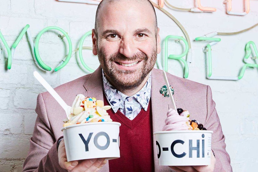 George Calombaris smiles at the camera holding two punnets of frozen yoghurt infront of a white wall with writing in lights.