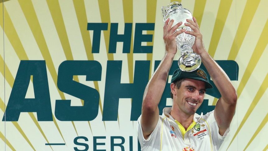 Australia cricket captain Pat Cummins lifts a crystal trophy shaped like the Ashes urn in front of a sign reading "THE ASHES".