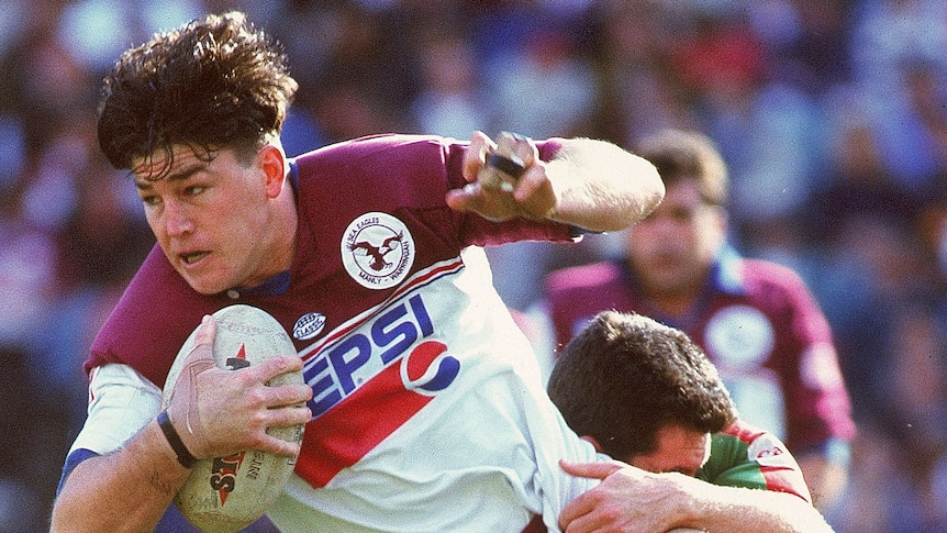 A Manly rugby league player holds the ball as he is tackled by a South Sydney opponent.