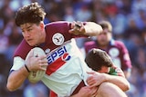 A Manly rugby league player holds the ball as he is tackled by a South Sydney opponent.