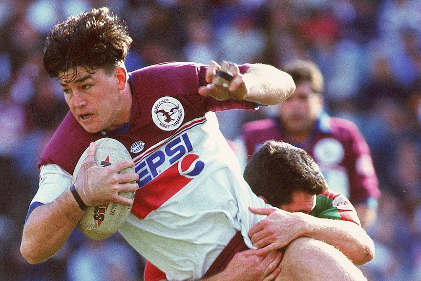 A Manly rugby league player holds the ball as he is tackled by a South Sydney opponent.