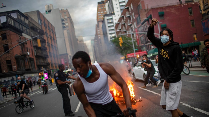 You look down a New York street with people protesting around debris on fire on an overcast day.