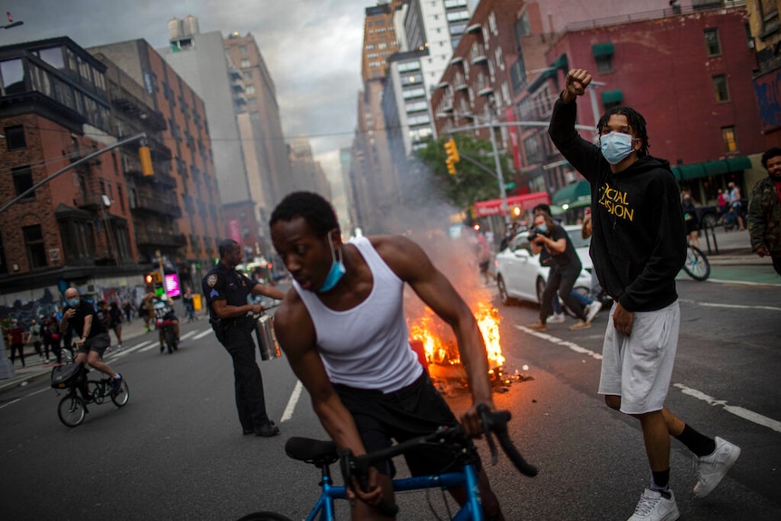 You look down a New York street with people protesting around debris on fire on an overcast day.