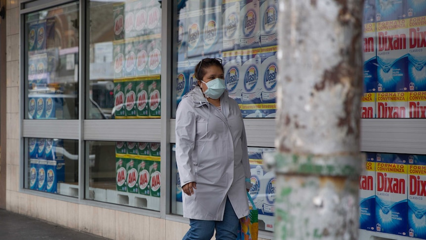 A woman wearing a face mask walks past a shop window filled with washing detergent.
