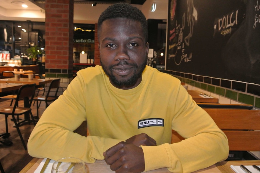 A young man in a yellow shirt smiles to camera, sitting down at a table in a cafe.