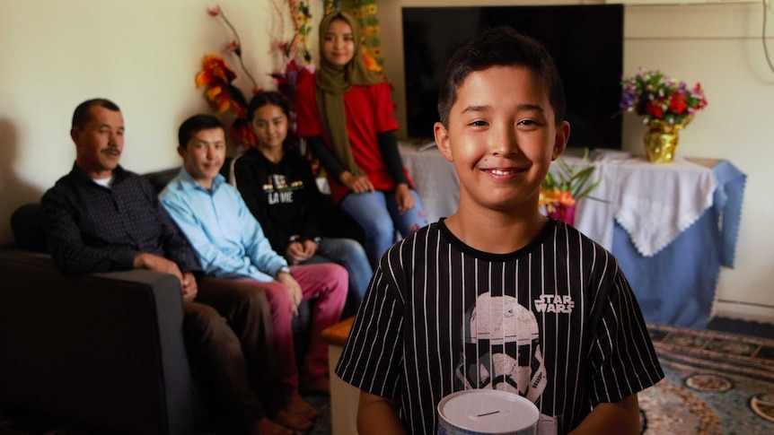 Murtaza Hussain stands holding a tin of pocket money with his family seated behind him