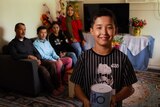 Murtaza Hussain stands holding a tin of pocket money with his family seated behind him