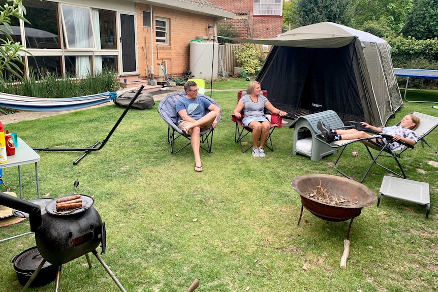 An man and woman sit with a young boy on camp chairs in the back garden with a tent and campfire pit