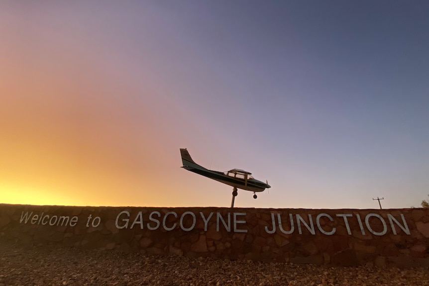 The silouete of a mounted light plane is above a sign saying "Welcome to Gascoyne Junction" in front of a sunrise sky. 