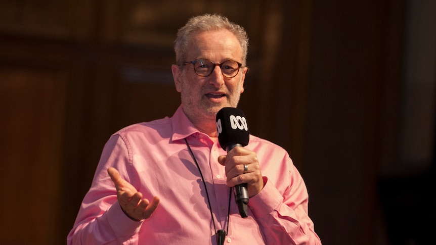 Jon Faine smiles in a pink shirt as he stands on stage in the Melbourne Town Hall holding an ABC microphone.