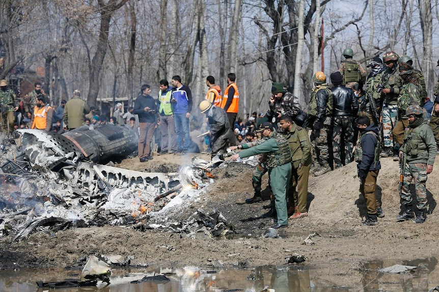 Military officials inspect the smouldering wreckage of a downed plane as a crowd of onlookers gather