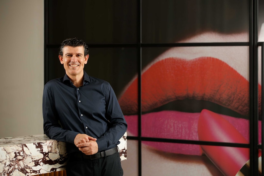A man stands in front of an add featuring lipstick being applied to lips.