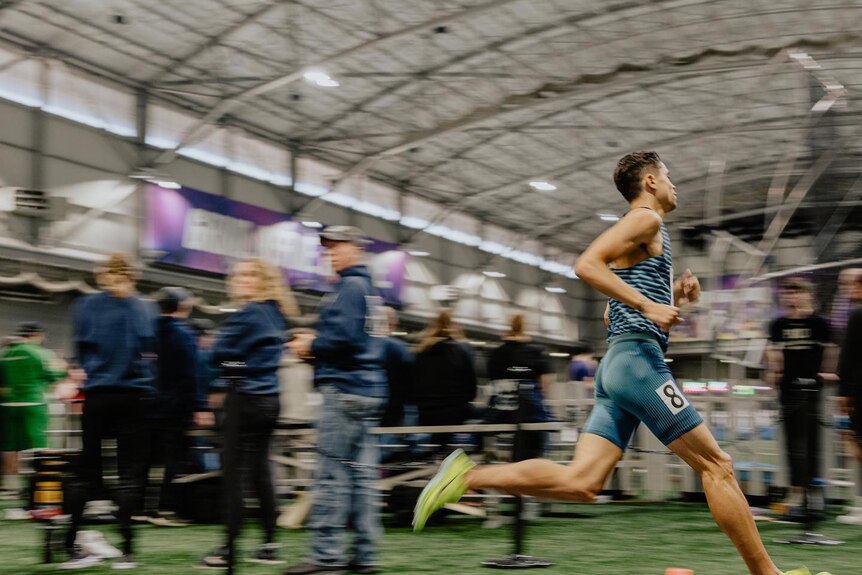 A man running on an indoor track, he is in focus, other people are blurred